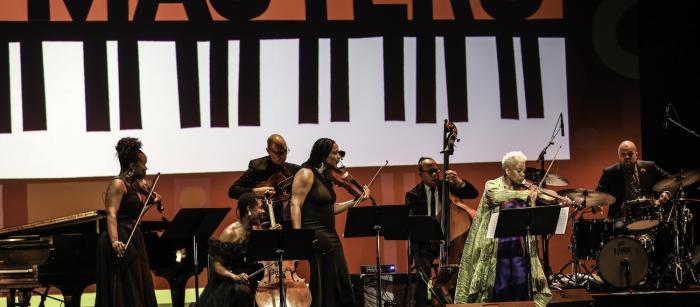 Regina Carter plays the violin at the front of the stage with a group of three other women holding violins and a cello watch her while a group of musicians perform behind them on the guitar, bass, and drums. The National Endowment for the Arts Jazz Masters logo is projected on the back wall. 