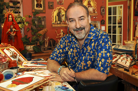 Artist in blue shirt patterned with gold crosses sitting in red-painted room, surrounded by his artwork of sculptures and painting.