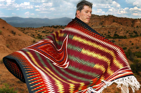 Man with woven rug around his shoulders standing in the middle of the desert. 