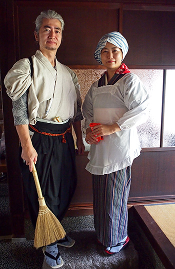 A man and woman in traditional Japanese dress hold small brooms