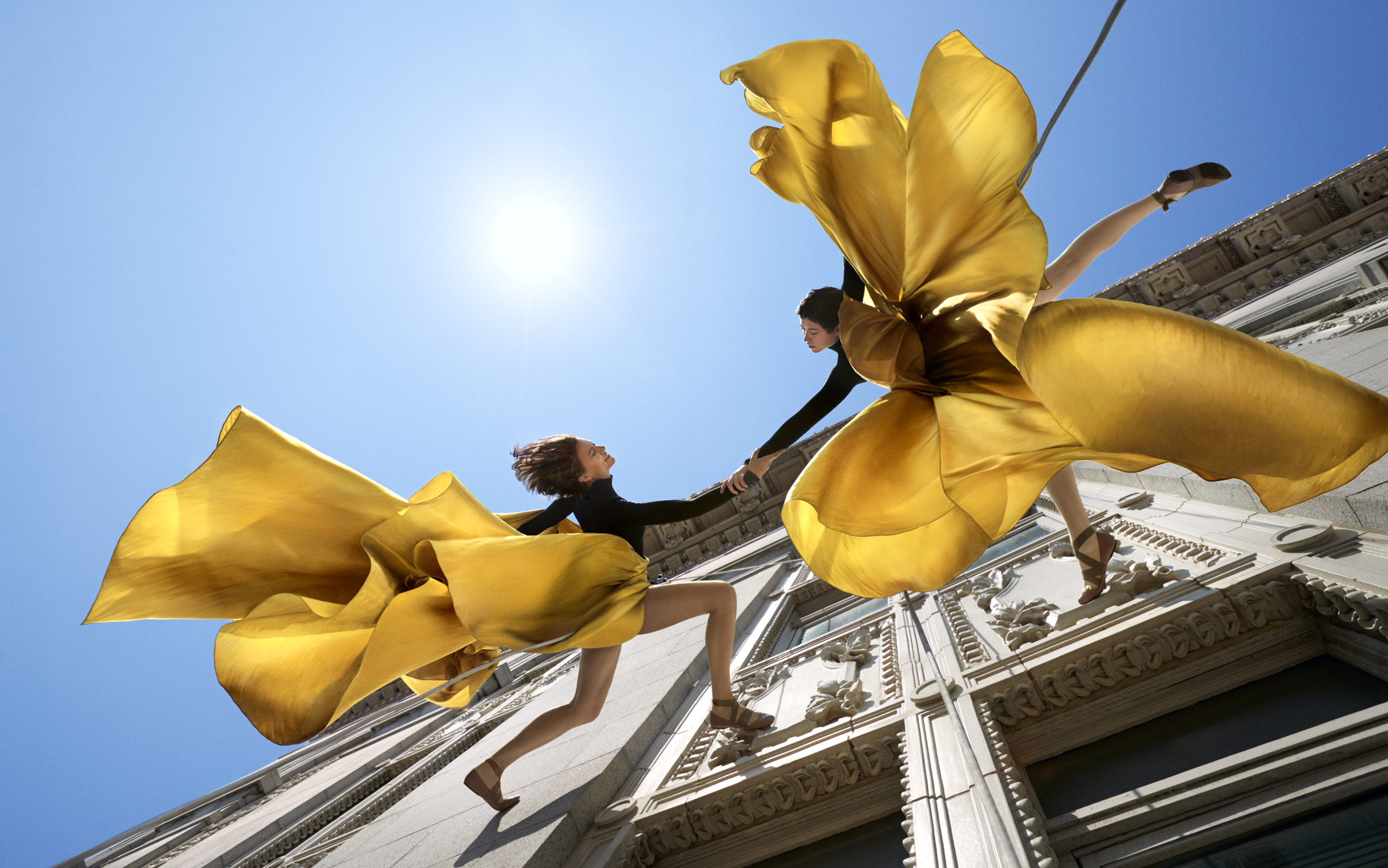Dancers with flying yellow skirts dance while suspended against a building wall