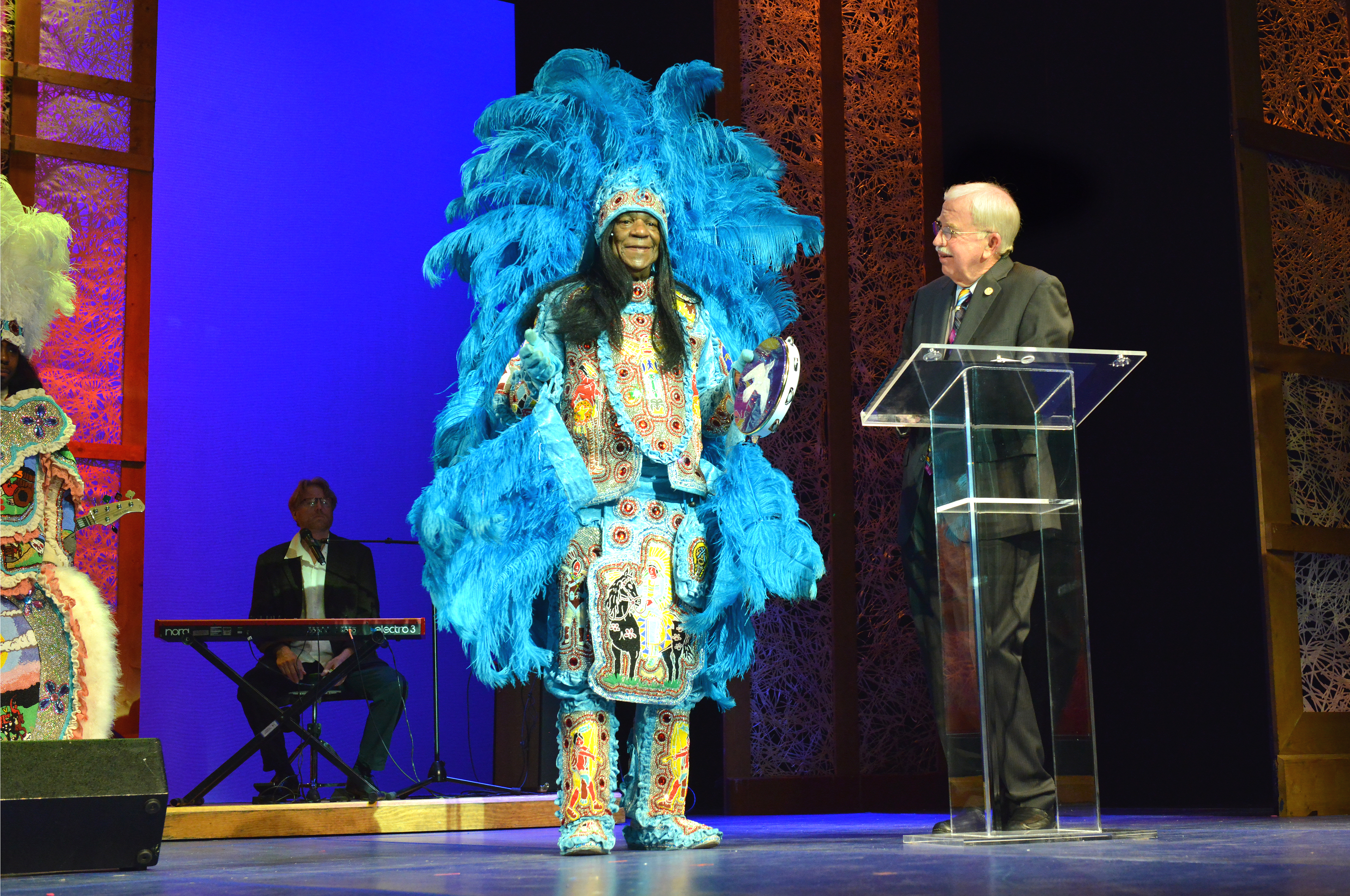 Honoree wearing elaborate custom with feathers and sequins. He is standing with emcee and talking. 