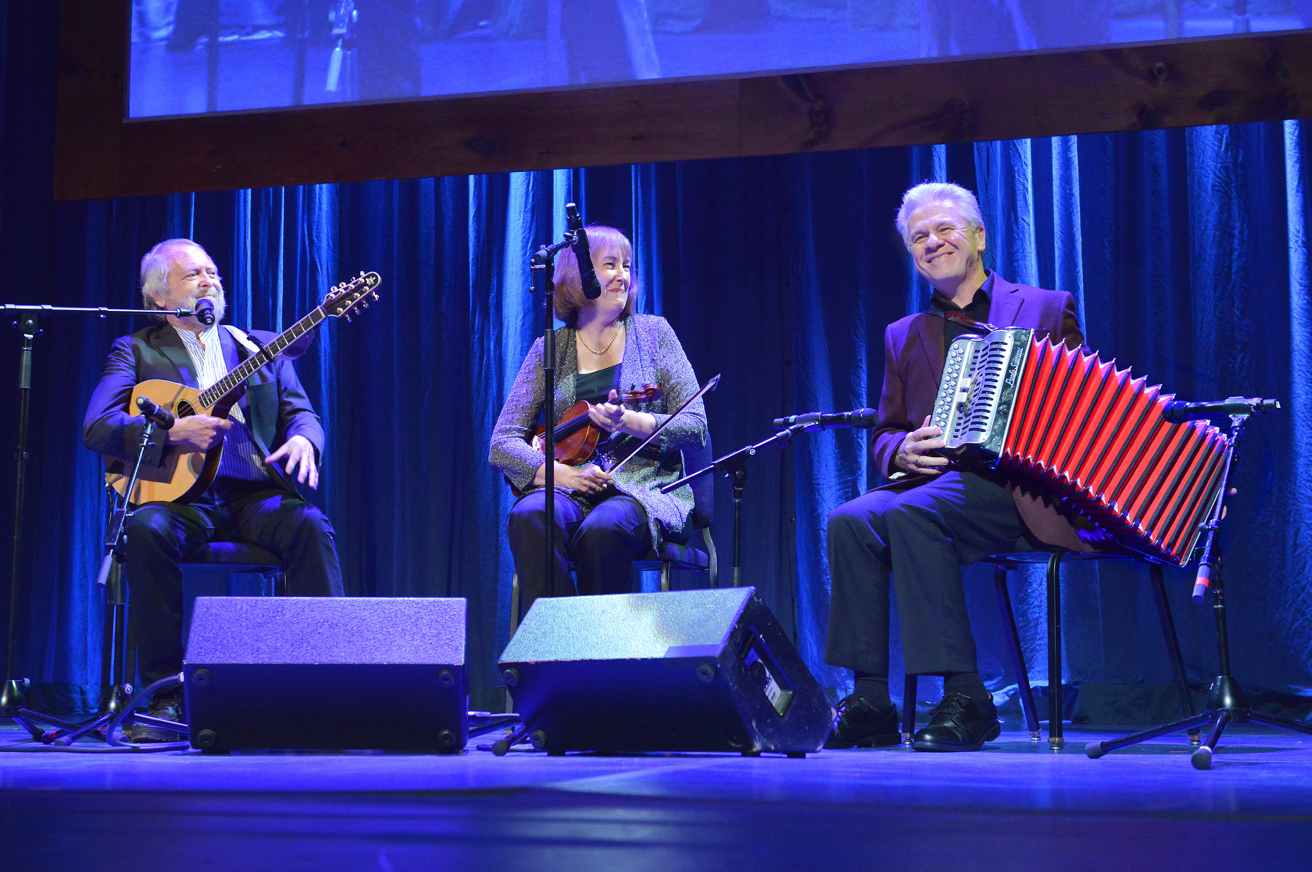 Three fellows sitting together on stage with instruments in their hands and playing music.
