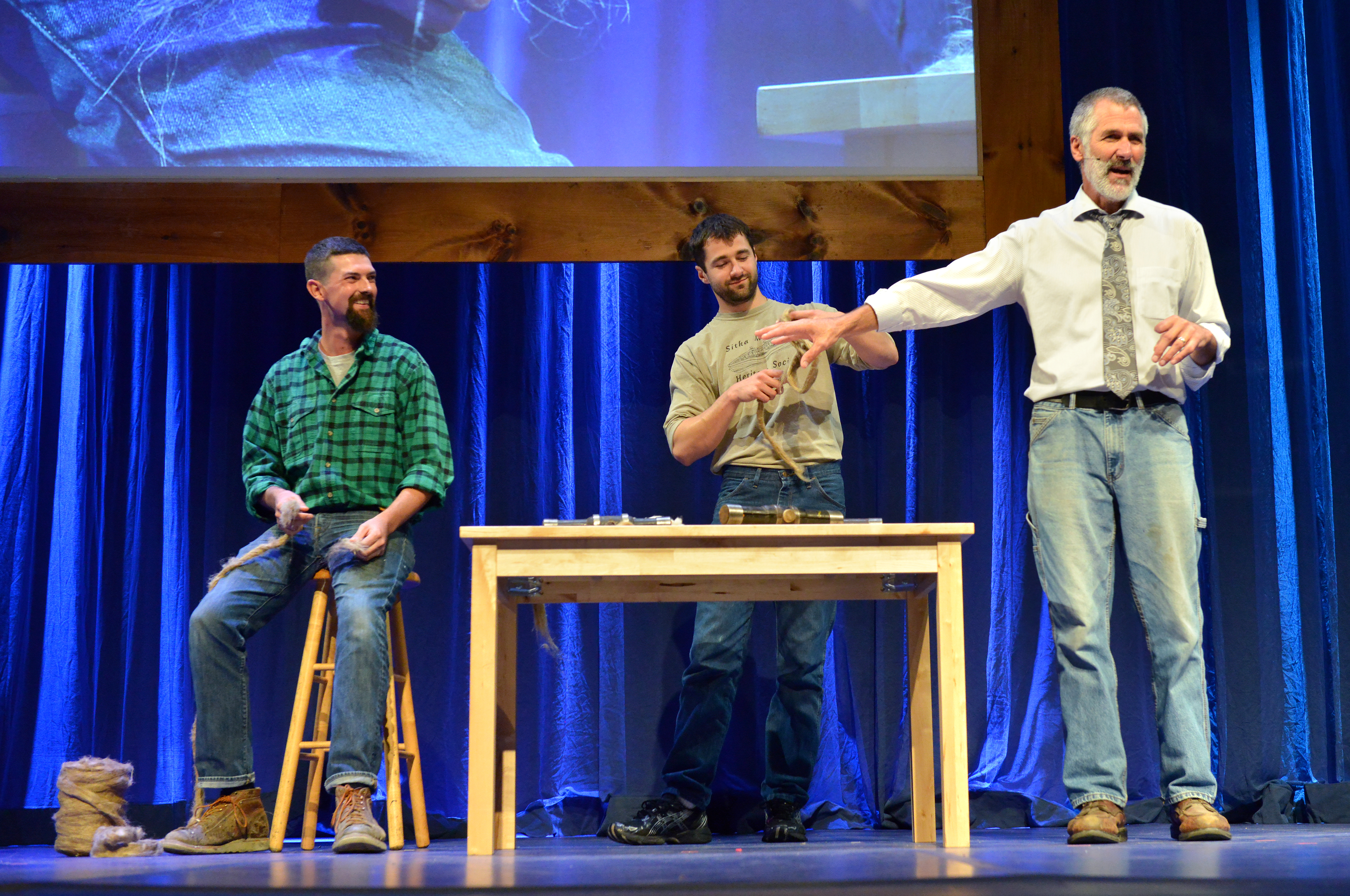 Two men on stage with shipbuilding materials in their hands while the fellow stands over them to explain what they are doing.