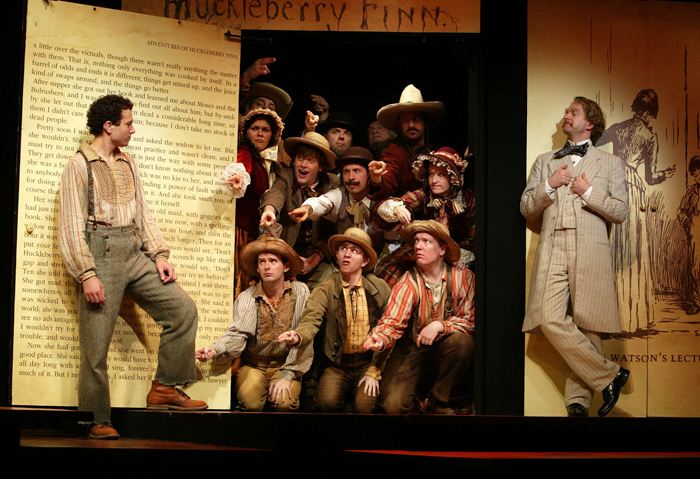 A cast dressed in 1800s clothing in front of a backdrop of Huckleberry Finn. 