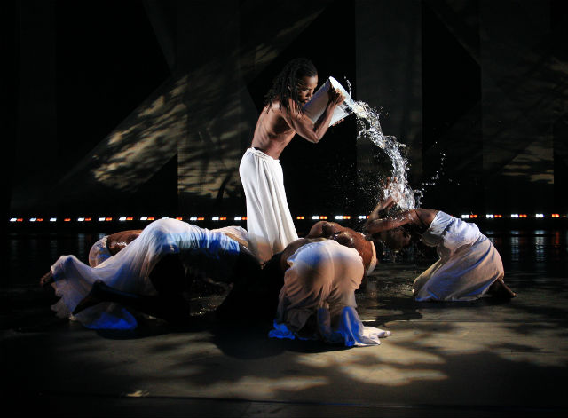 A dancer pours a bucket of water over other crouching dancers in a performance