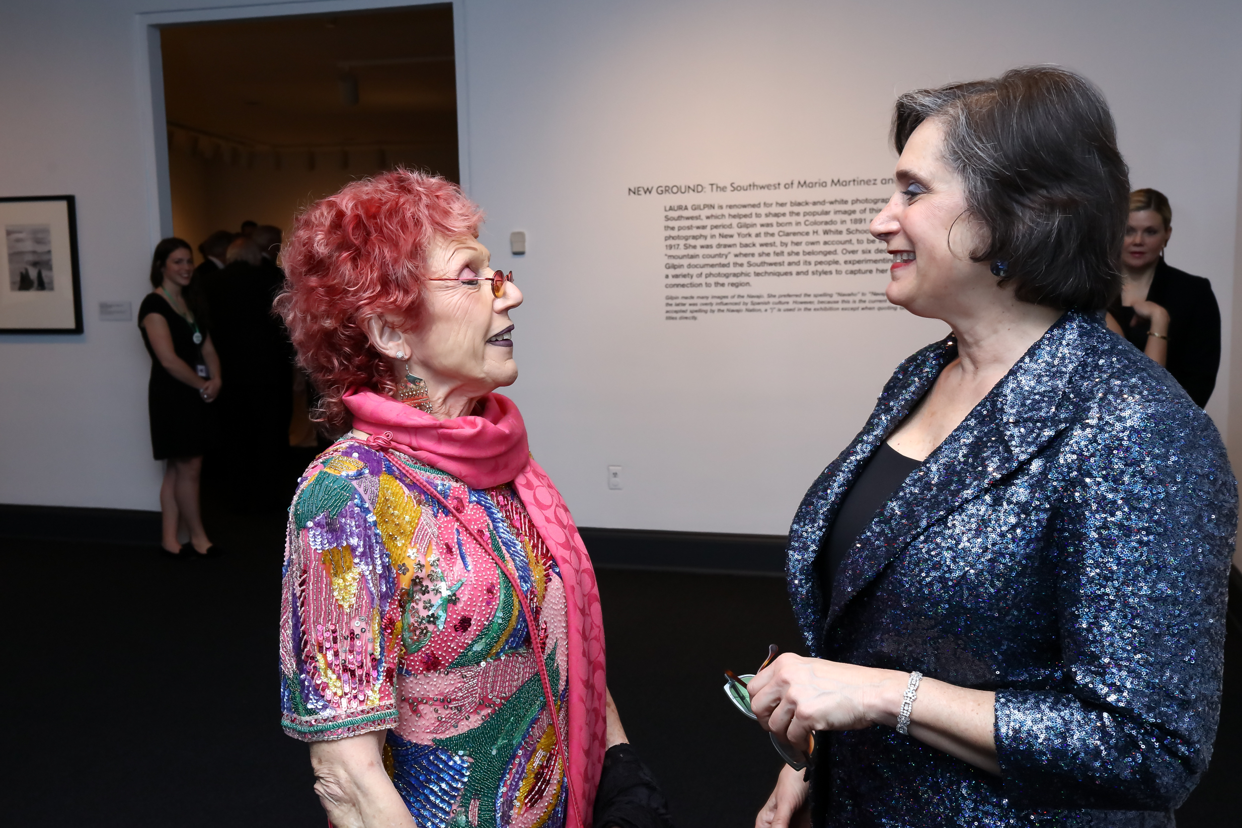 Woman with bright red hair and colorful dress talking to another woman in blue sparkly blazer in a museum.