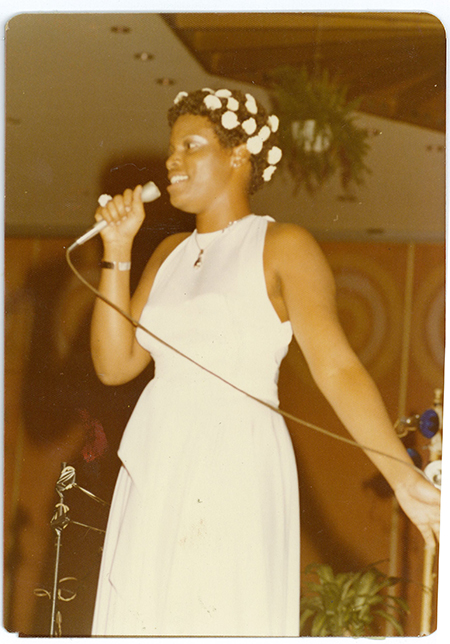 Girl in white dress with flowers in her hair singing with arm outstretched.