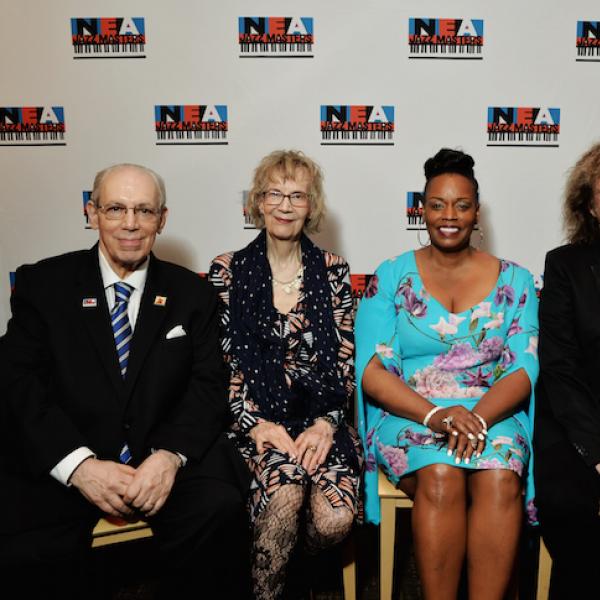 Two men and two women sit in chairs in front of a banner with the NEA Jazz Masters logo repeated.