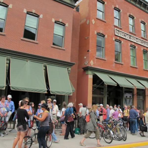 People standing in a line in front of old brick buildings with green awnings to get tickets to a movie.