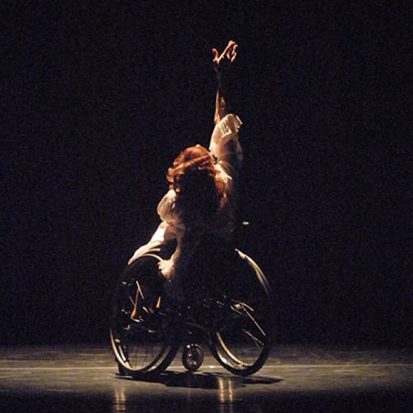 A dancer in a wheelchair lifts one hand upward as she performs onstage