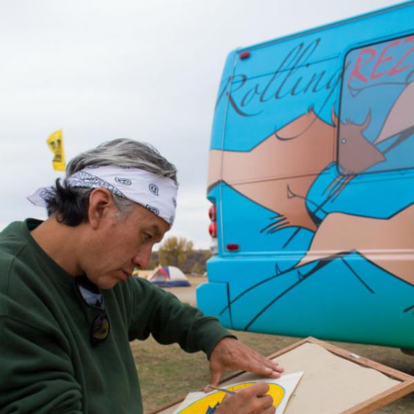 A man in bandana does a watercolor painting in front of a colorful bus