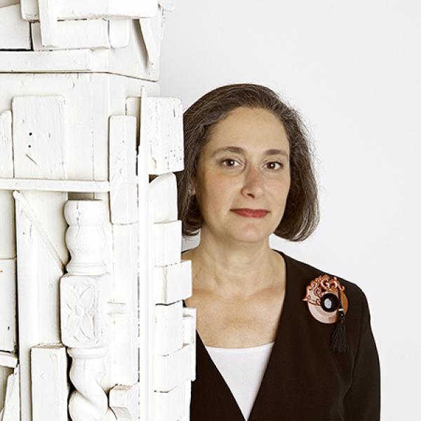 Woman wearing a black suit with large red brooch standing next to a white sculpture. 