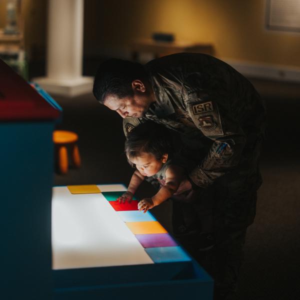 A father dressed in a military uniform holds his young son at an interactive art exhibit. The background is dark and there is light shining up illuminating their faces