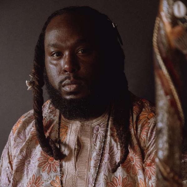 Portrait of Black man with beard and long braided hair wearing a orange flowered shirt and holding a staff.