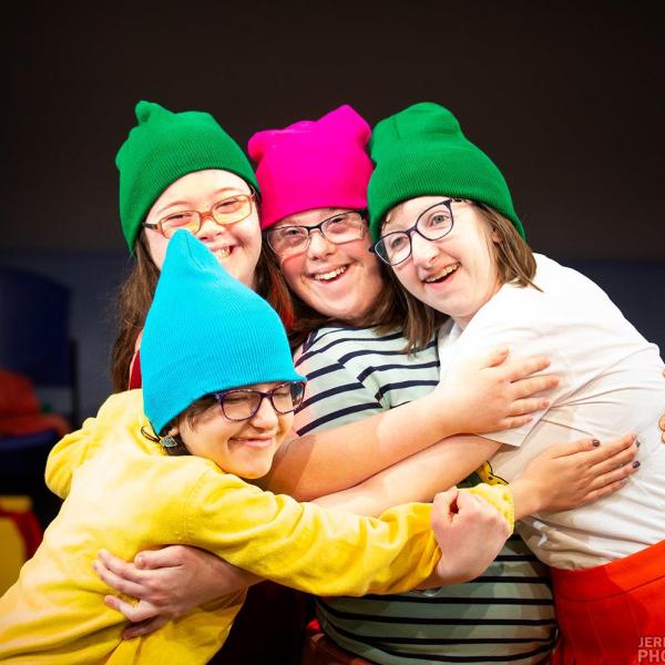 Four young actors on stage embracing each other and wearing glasses and a range of colorful hats (green, pink, and blue).