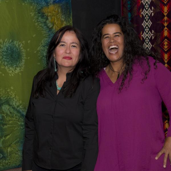 Two women in front of artwork, one dressed in black with long black hair and not smiling, the other dressed in purple and laughing.