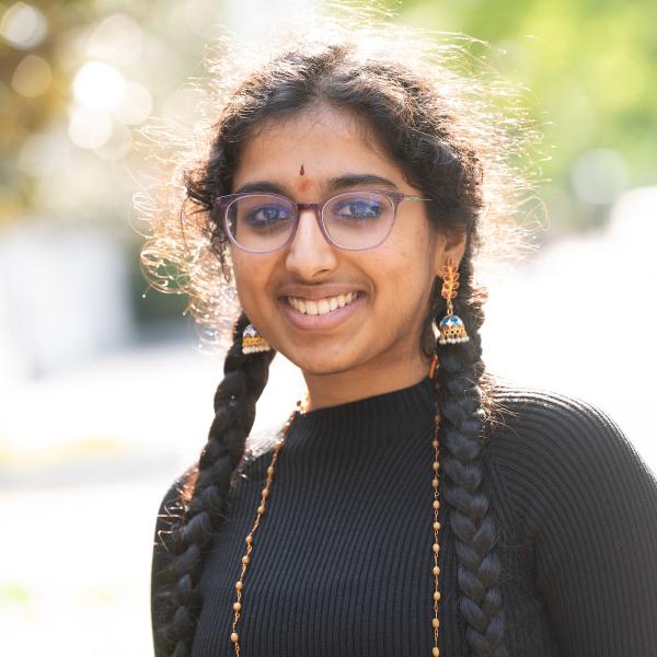 A young South Asian woman smiles, wearing a black turtleneck, long necklace, and dangling earrings. She has glasses and two long braids.