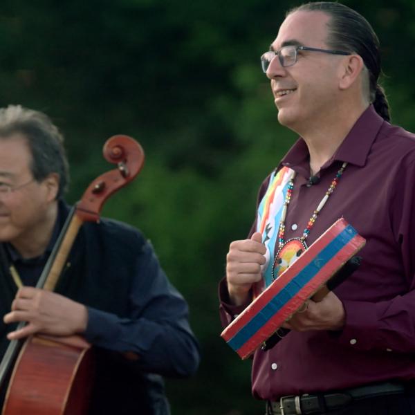 An Asian man in black shirt playing a cello next to a Native man in a purple shirt playing a drum.