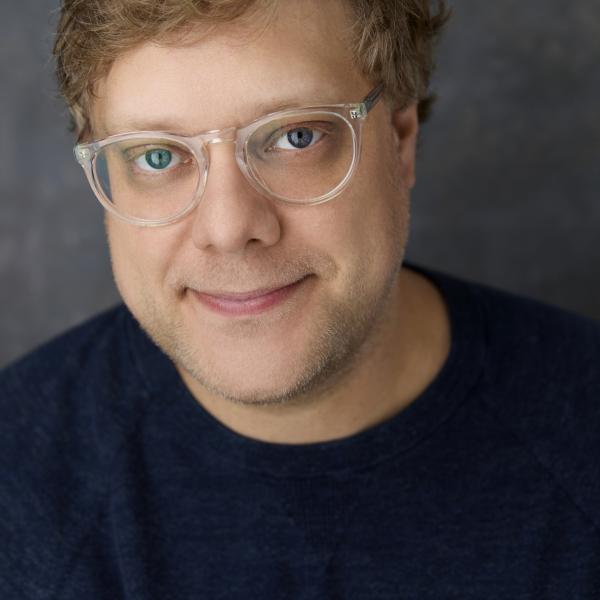 Headshot of Andrew Dolan, a white man with sandy blonde hair, glasses, and a blue eyes. He wears a grey shirt. 