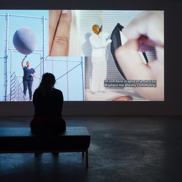 Woman sitting on a bench in a dark room, facing an art exhibit that depicts a woman holding a large ball and a painting of a black man in a white suit, with text in the exhibit that says: "I'm still here in spite of efforts to displace me and my community."