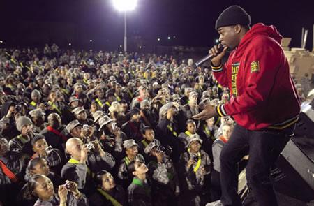 African-American man in red sweat jacket and wearing black hat rapping to a crowd of servicemen.