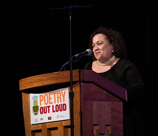 A woman speaks at a podium with a Poetry Out Loud banner hanging from it