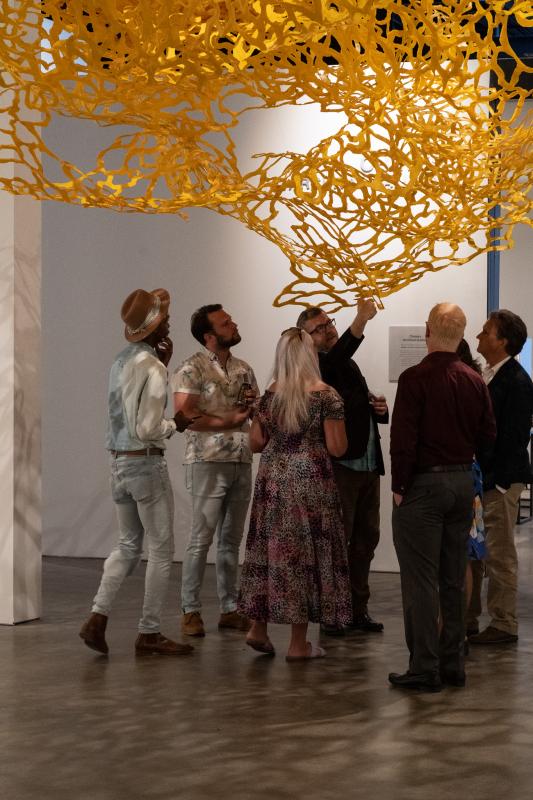 A group of people look up in a gallery at a lacy yellow sculpture installation