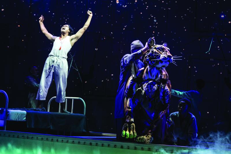 On stage, man in white shirt and pants at left with arms in the air shouting; to the right two people behind a paper mache tiger. 