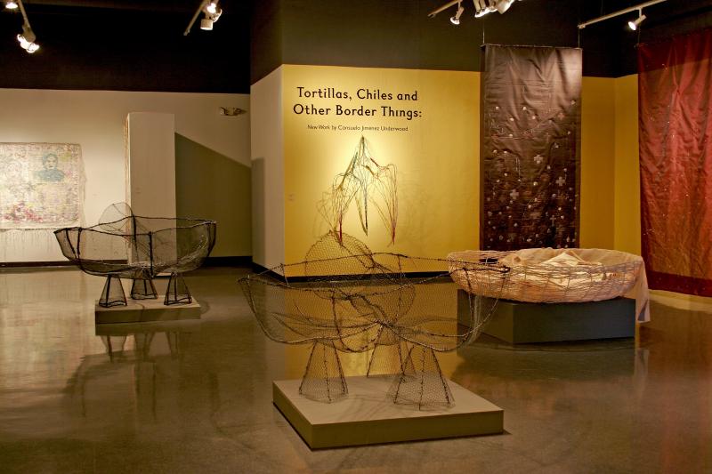 Exhibition space with brown and tan woven artwork on display. The wall in the middle of the exhibit says "Tortillas, Chiles and Other Border Things"