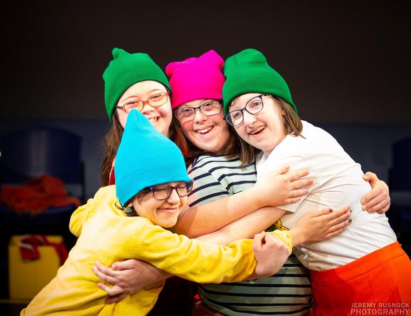 Four young actors on stage embracing each other and wearing glasses and a range of colorful hats (green, pink, and blue).