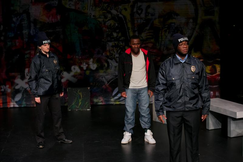 On stage: White actress (left) in a black police outfit glaring at a Black actor (center) wearing jeans and a black and pink hoodie, who is glaring at a Black actor (right) who is wearing glasses and a black police outfit.