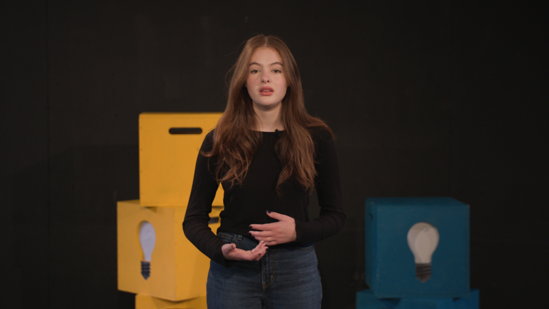 A young white woman recites in front of stacked boxes and a black background