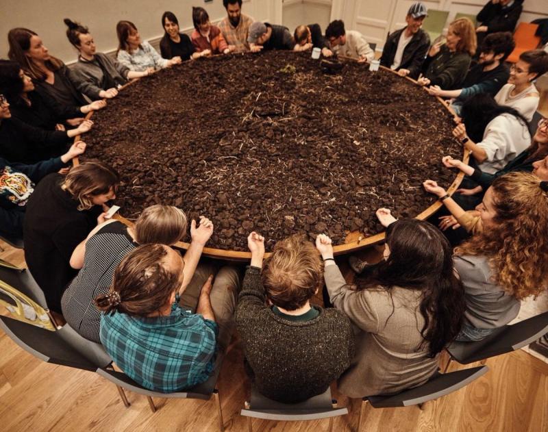 A group of people sit around a large table with their hands outstretched, feeling a large pile of dirt that covers the surface.