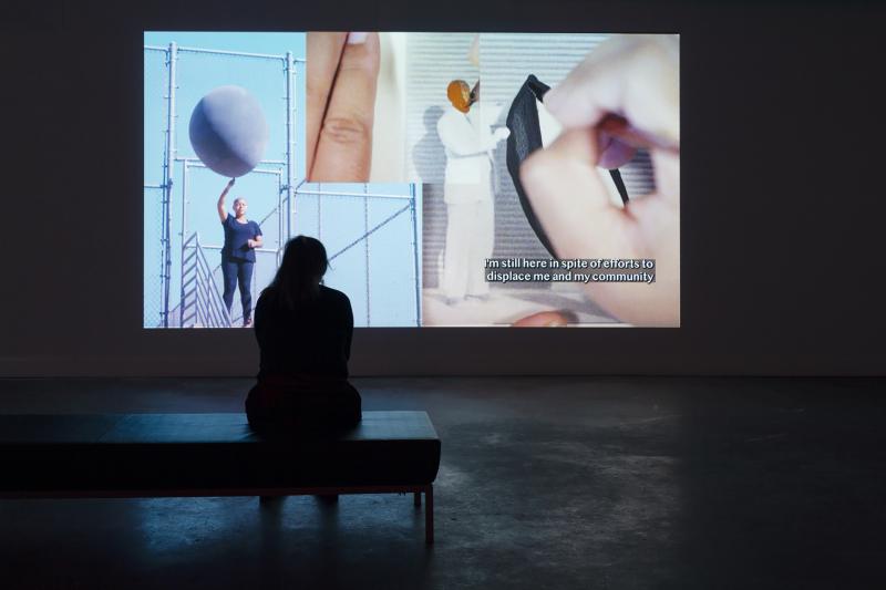 Woman sitting on a bench in a dark room, facing an art exhibit (digital video) that depicts a woman holding a large ball and a painting of a black man in a white suit, with text in the exhibit that says: "I'm still here in spite of efforts to displace me and my community."