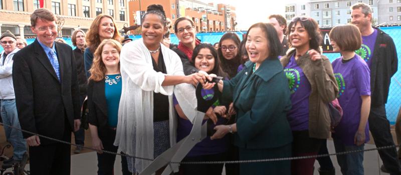 Group of people clustered together around a woman with a large pair of scissors ready to cut the ribbon to open an event