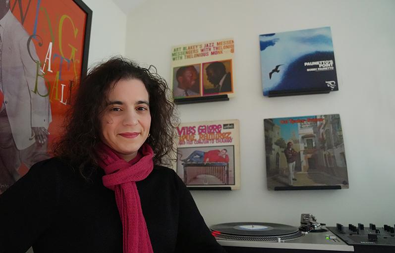 a white woman with long curly dark hair sits with a record player and framed albums behind her