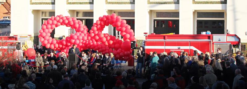 Large crowd at an opening event with a speaker at the podium, celebratory balloons, fire engines