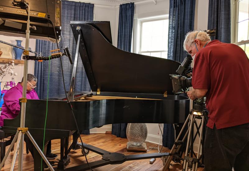 Black woman (wearing a purple shirt) on a film set playing a piano. A White man (wearing a mask and red shirt) is standing and filming the Black woman.