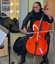 Juleaka Brown, a Black woman with braids sits playing a cello