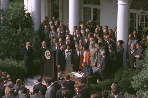 Man in suit sitting at table outside the White House surrounded by many other men in suits and a woman in an orange jacket. 