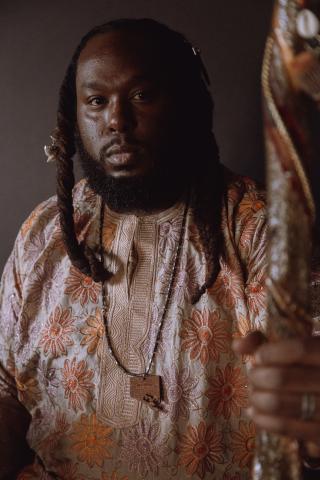 Portrait of Black man with beard and long braided hair wearing a orange flowered shirt and holding a staff.