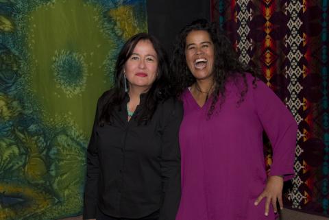 Two women in front of artwork, one dressed in black with long black hair and not smiling, the other dressed in purple and laughing.