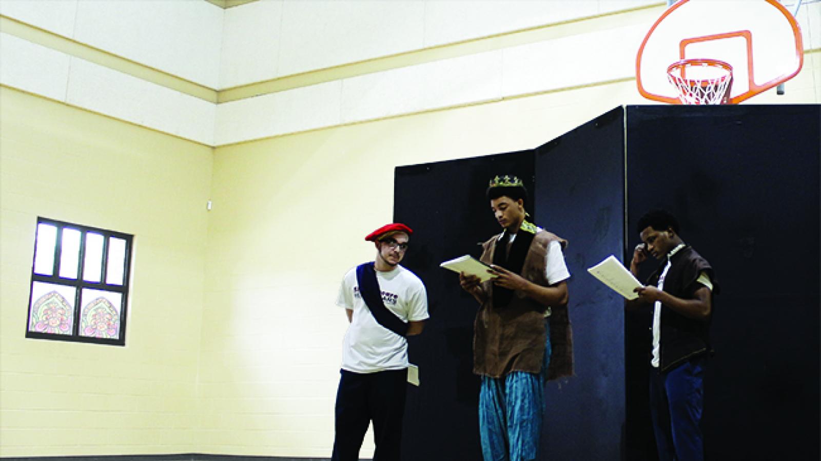 Three young men wearing costumes read from scripts in a gymnasium