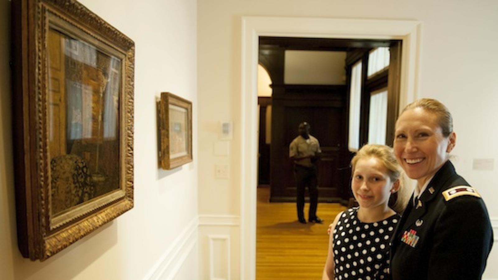 A white woman in military dress stands in front of two paintings with her young daughter