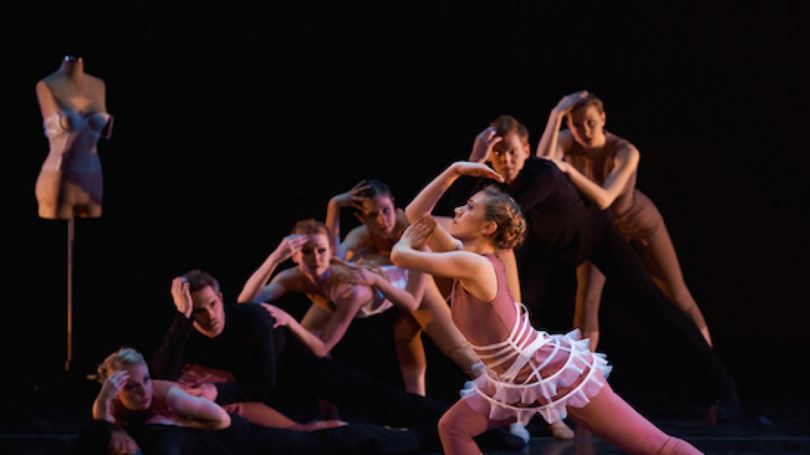 A female dancer in profile leans toward the left with her hand bent on her head while several dancers behind her mimic the move
