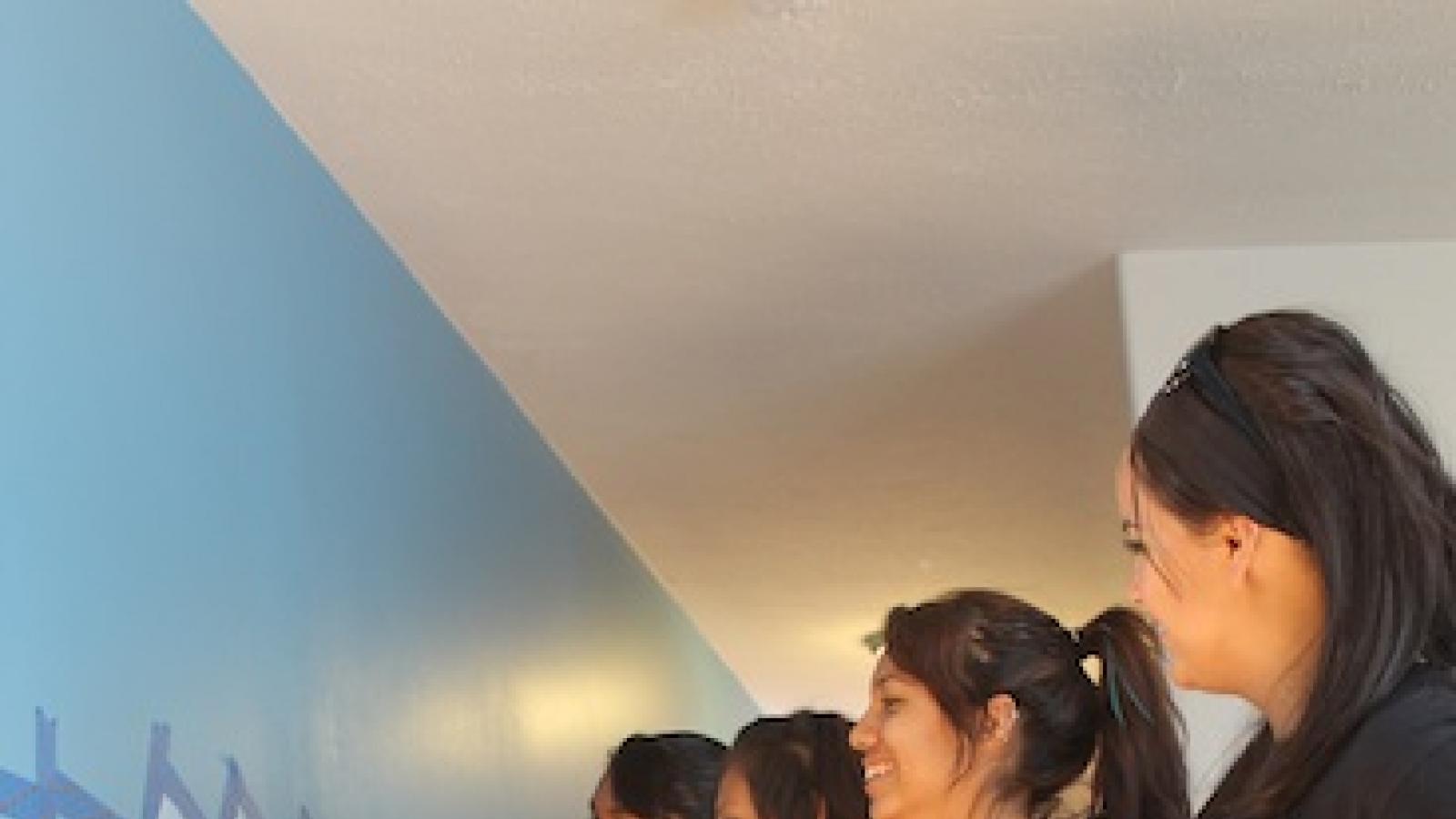Four young Native American girls in casual clothing paint a mural in blue, yellow, and orange on an office wall