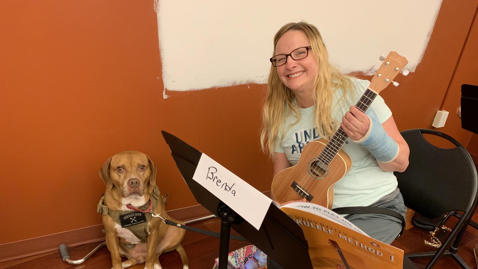 Woman with ukelele sitting and smiling with service dog sitting next to her.