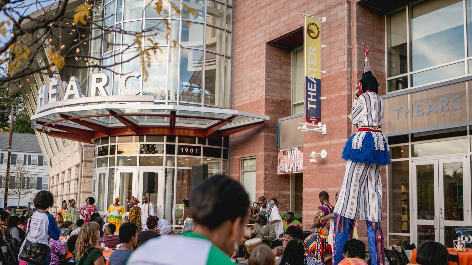 A crowd outside a glass-fronted building, and a person on stilts walking through the crowd. 