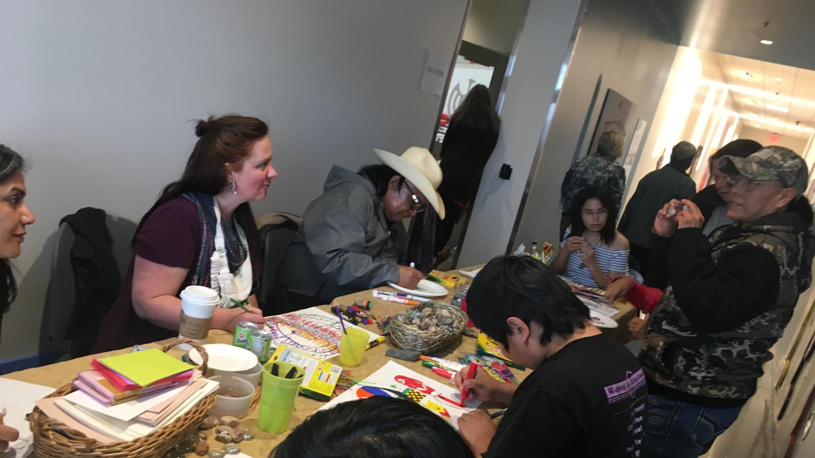 A group of people sitting around a long table with art supplies on it. 